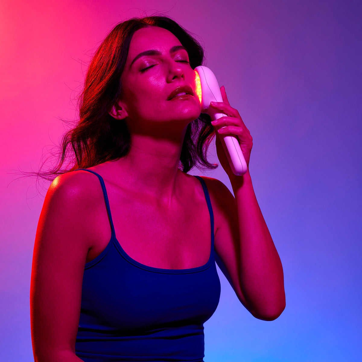 A woman with her eyes closed holds a white handheld beauty device against her face, illuminated with a pink and yellow light, set against a vibrant pink and blue gradient background.