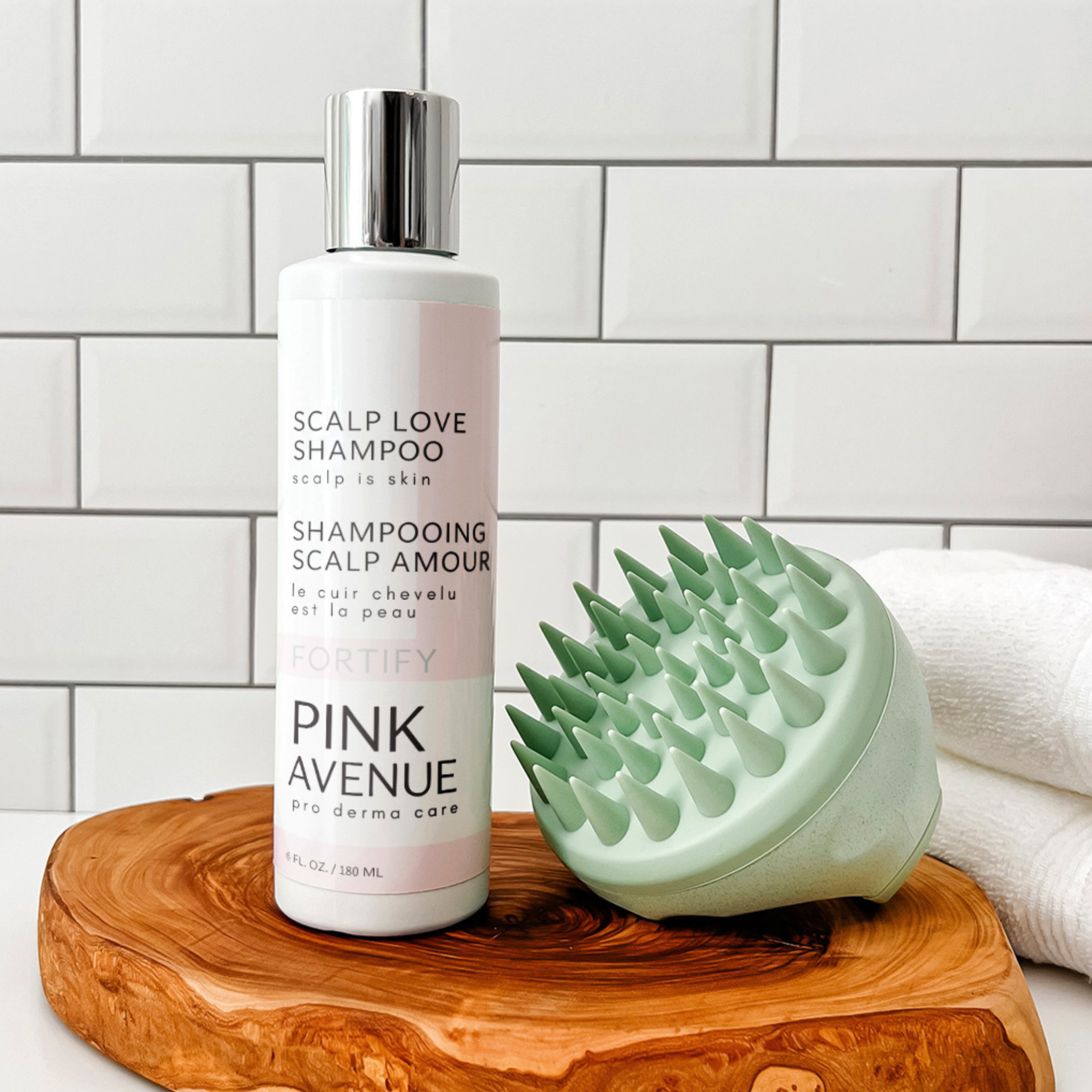 Pink Avenue Shampoo bottle and scalp massager on a wooden stand with a tiled wall background